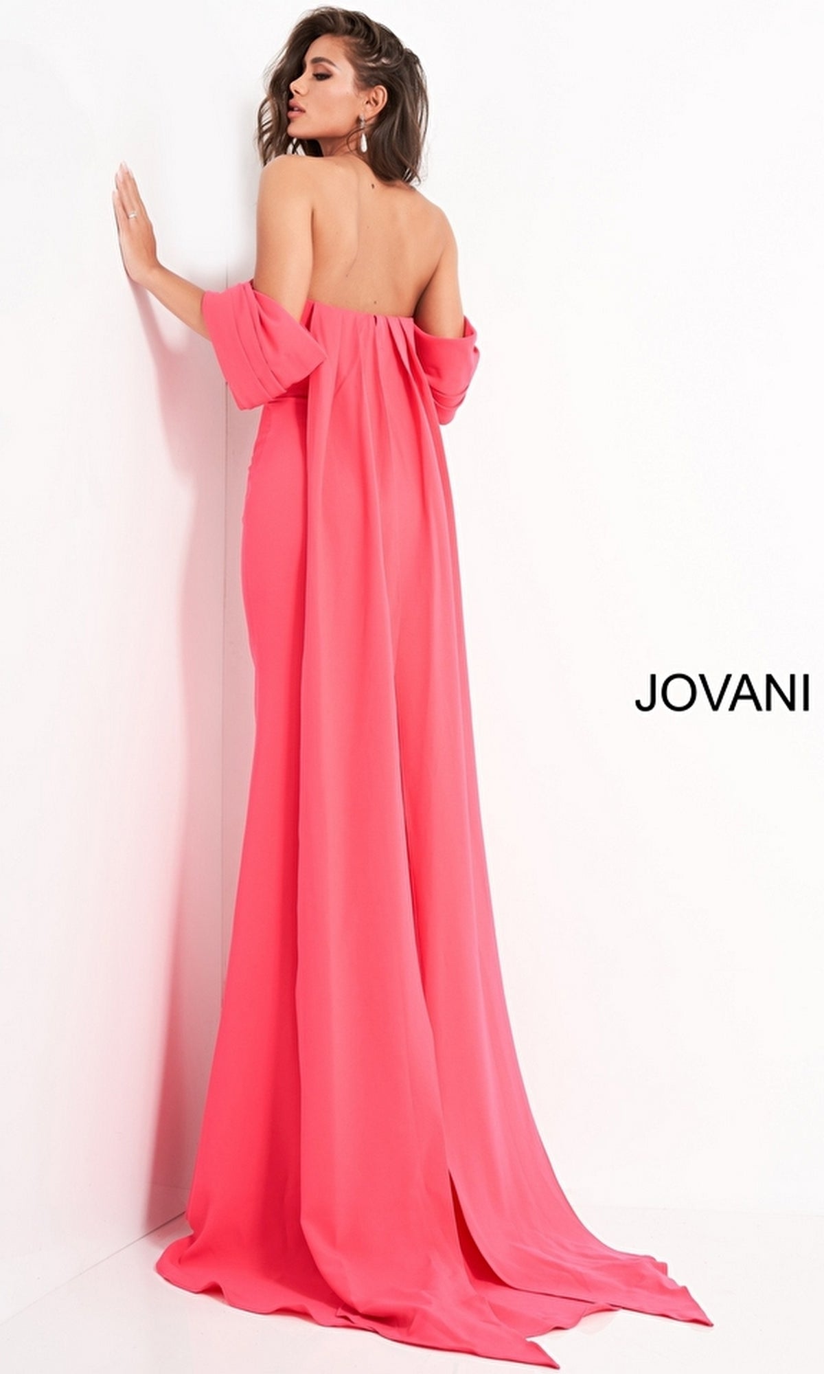 A woman stands against a white wall wearing the Long Prom Dress by Jovani 04350, a strapless, floor-length pink gown with a long train. The word